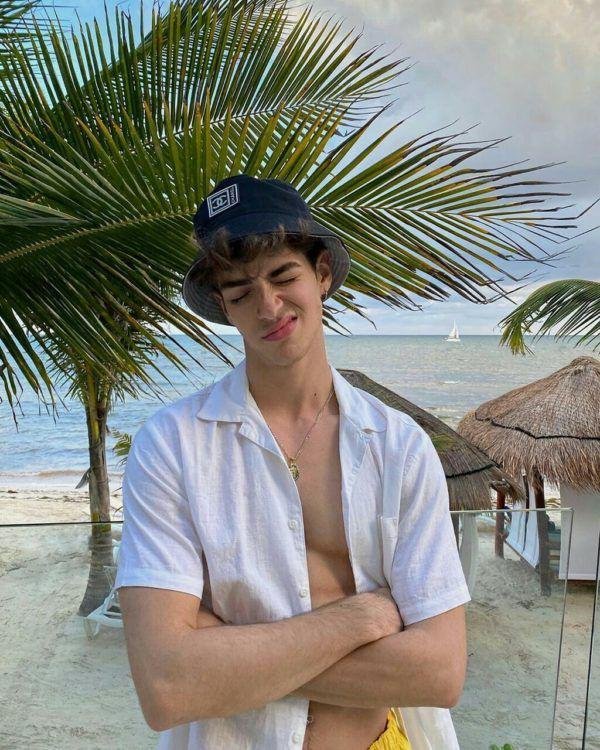 Chico con los brazos cruzados y camisa abierta junto a una palmera