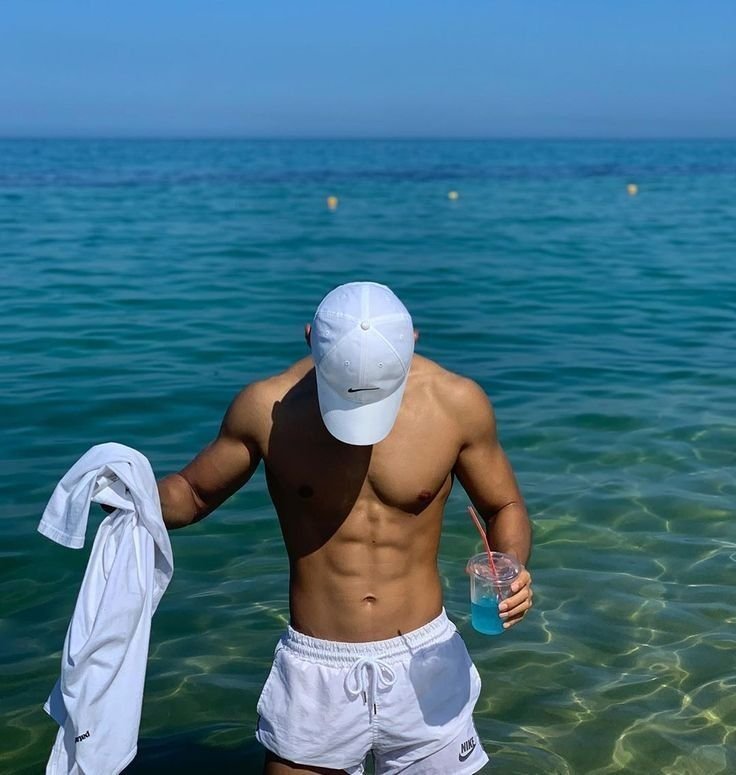 Chico de pie en el agua con gorra blanca y bebida